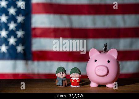 Pinkfarbene Schweinebank mit älteren Miniaturen auf einem Holztisch vor einer verschwommenen US-Flagge. Steht für amerikanische Altersvorsorge, Sozialversicherung, Stockfoto