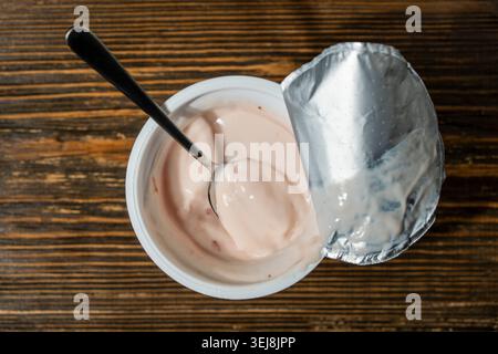 Blick von oben auf offenen Erdbeeren- oder Beerenjoghurt in einem Plastikbecher mit einem Metalllöffel innen. Serviert auf einem dunklen rustikalen Holztisch. Konzept für gesundes Brea Stockfoto