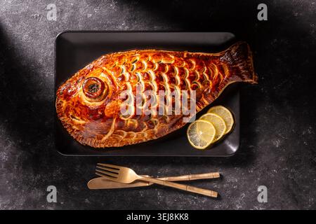 Gebackener Fischkuchen in Blätterteig, der wie ein Fisch geformt ist. Blick von oben auf traditionellen goldenen Fisch en Croute Pie serviert mit Zitronenscheiben auf einem schwarzen Teller. Kreativ Stockfoto