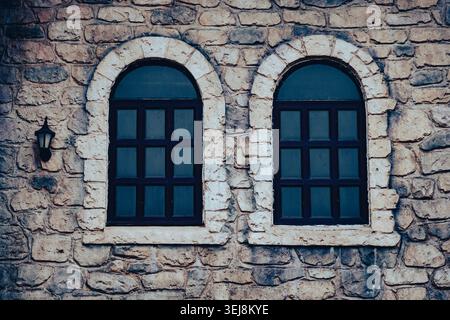 Zwei alte Bogenfenster an einer rustikalen Steinmauer eines alten Hauses. Symmetrische Aufnahme von zwei klassischen Bogenfenstern mit dunklen Rahmen, die in eine ancie eingebaut sind Stockfoto