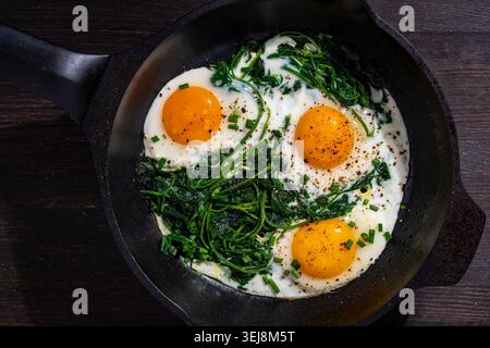 Spiegeleier mit Gemüse in einer Pfanne, Nahaufnahme. Blick von oben auf drei sonnige Spiegeleier, die mit frischem grünem Spinat und Gewürzen in einer schwarzen Pfanne auf einem Tag zubereitet werden Stockfoto