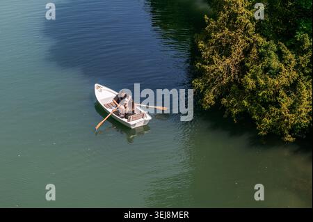Augsburg, Deutschland - 25. Mai 2026: Ein Hochwinkelblick eines Paares, das ein kleines weißes Boot mit Holzrudern auf einem ruhigen Fluss neben üppig grünen Bäumen rudert. Stockfoto