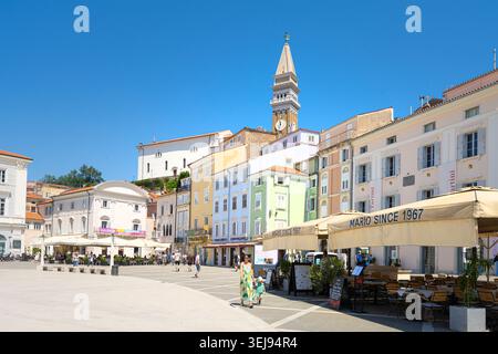 Piran, Slowenien. Mai 2026. Ein sonniger Tartini-Platz ist gesäumt von farbenfrohen Gebäuden und einem markanten Glockenturm. Man sieht Menschen beim Gehen und Essen Stockfoto