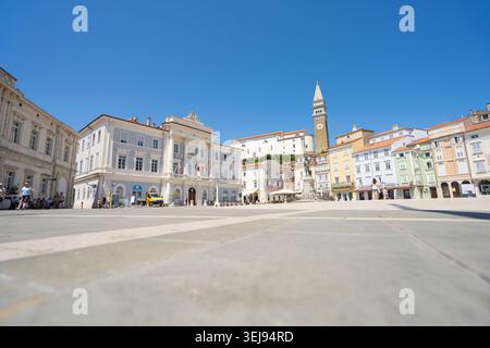Piran, Slowenien. Mai 2026. Ein sonniger Tartini-Platz ist gesäumt von farbenfrohen Gebäuden und einem markanten Glockenturm. Man sieht Menschen beim Gehen und Essen Stockfoto