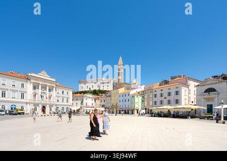 Piran, Slowenien. Mai 2026. Ein sonniger Tartini-Platz ist gesäumt von farbenfrohen Gebäuden und einem markanten Glockenturm. Man sieht Menschen beim Gehen und beim Essen Stockfoto