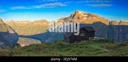 Ljøen, Norwegen - 24.05.2026: Ein Weitwinkel-Landschaftsfoto einer dunklen Holzhütte auf einem üppigen, grasbewachsenen Hügel in Ljøen, Norwegen, mit Blick auf einen tiefen bl Stockfoto