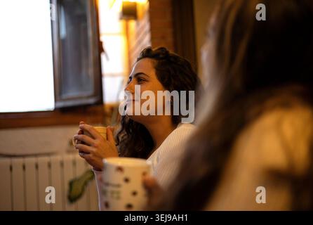 Junge Frau lächelt, hält heiße Kaffeetasse, während sie sich mit einem Freund unterhalten Stockfoto