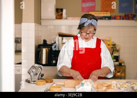 Ältere kaukasische Frau in roter Schürze, die in einer hellen professionellen Küchenwerkstatt handwerklichen Teig zubereitete Stockfoto
