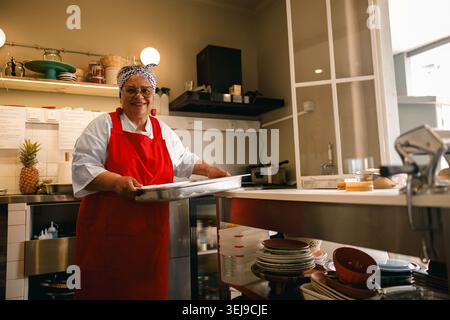 Lächelnde hispanische Seniorin, die ein Backblech in einem professionellen Restaurant hält Stockfoto