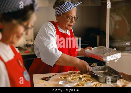 Ranghohe Afroamerikanerin, die in einer Gemeinschaftsküche Essen zubereitet, trägt rote Schürze und Stirnband. Stockfoto