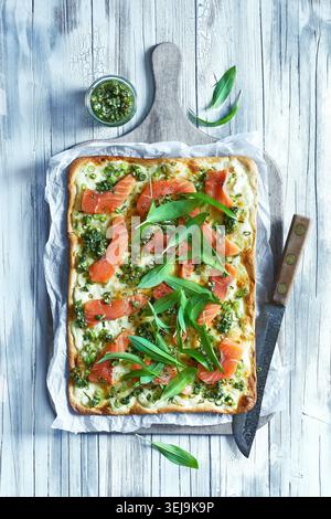Rustikale Fotos von einem frisch gebackenen Flamkuchen mit Räucherlachs, wilden Knoblauchblättern und cremigem Käse. Das Fladenbrot ruht auf Parchm Stockfoto