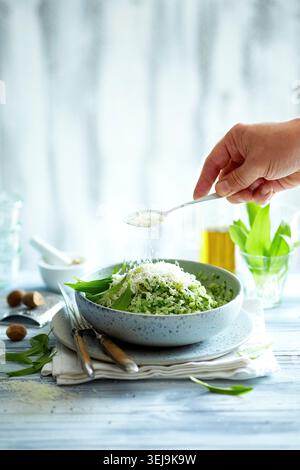 Hochwertiges, redaktionelles Foto von einem cremigen wilden Knoblauchrisotto, serviert in einer rustikalen Keramikschale. Eine Hand streut frisch geriebenen Parmesan aus einem Sp Stockfoto