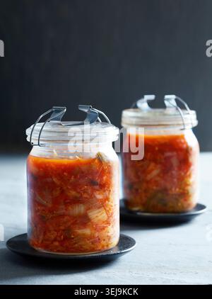 Zwei Glasgläser gefüllt mit hausgemachten Kimchi, platziert auf dunklen Keramikplatten vor einem stimmungsvollen Hintergrund. Die lebhafte, rotfermentierte napa-Kohl-Mischung, Stockfoto