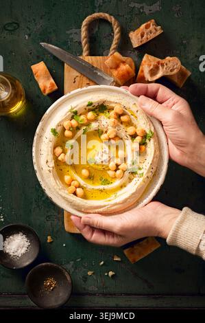 Flache Lage mit frischem hausgemachtem Hummus-Dip auf grünem Hintergrund, mit frischen Kichererbsen und Petersilie, Olivenöl und Pitabrot gestylt, hält eine Frau das Stockfoto