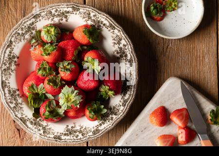 Frische Erdbeeren in einer Vintage-Schüssel mit geschnittenen Beeren auf einem Brett Stockfoto