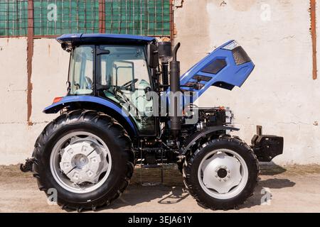 Ein blauer Nutztraktor mit offener Motorhaube steht bei Wartungsarbeiten im Betrieb im hellen Sonnenlicht Stockfoto