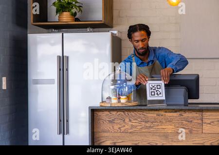 Afroamerikanischer Mann in grüner Schürze, das QR-Zeichen an der Kasse mit Cupcakes, Kopierraum Stockfoto