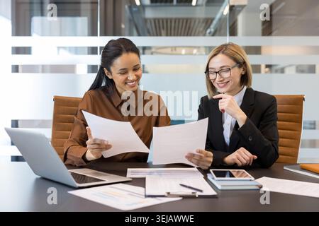 Zwei lächelnde Geschäftsfrauen sitzen an einem Bürotisch, prüfen Finanzdokumente und diskutieren ein Arbeitsprojekt, während sie in einer professionellen Unternehmensumgebung zusammenarbeiten Stockfoto