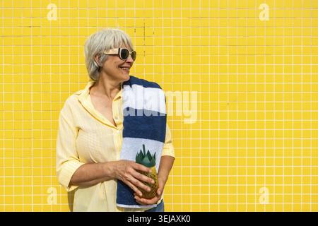 Ältere Frau mit grauen Haaren lächelt, trägt eine Sonnenbrille, hält ein gestreiftes Handtuch und eine ananasförmige Flasche an einer hellgelben gefliesten Wand, Emb Stockfoto