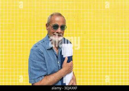 Ein älterer Mann mit grauem Bart und runder grüner Sonnenbrille lächelt fröhlich, hält ein gestreiftes Strandtuch über den Arm und steht gegen ein helles Geschrei Stockfoto