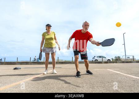Seniorenpaar, die einen aktiven Lebensstil genießen, indem sie an einem sonnigen Tag in der Nähe der Küste Pickleball spielen und dabei auf gesundes Altern, Fitness und Ruhestand hinweisen Stockfoto