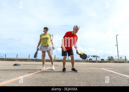 Aktives Seniorenpaar, das auf einem Asphaltplatz am Meer pickleball spielt und Fitness, Gesellschaft und Outdoor-Erholung während einer sonnigen, heiklen Jahreszeit genießt Stockfoto