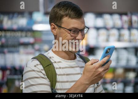 Mann mit Brille und Rucksack glücklich überrascht und lächelt, als er das Smartphone im Laden ansieht und etwas auf dem Bildschirm ansieht. Der Kunde ist online zufrieden Stockfoto