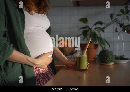 Schwangere Frau, die ihren Bauch hält, in einer Küche steht und einen lebendigen grünen Smoothie mit einem Strohhalm hält, der sich auf gesunde Ernährung, Vitamine, und konzentriert Stockfoto