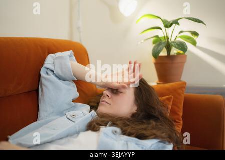 Junge Frau, die unter schmerzhaften Kopfschmerzen oder Migräne leidet, zu Hause auf einem orangen Sofa liegt, sich gestresst fühlt, unwohl ist und eine Krankheit einsetzt Stockfoto