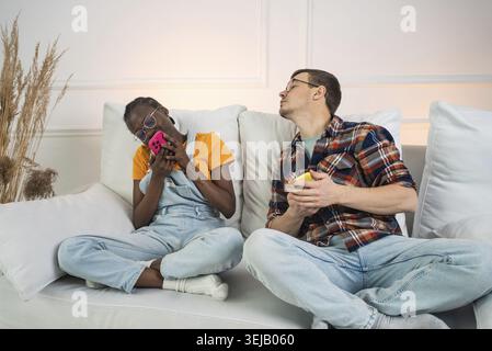 Multiethnische Paare sitzen auf einem weißen Sofa, ignorieren sich gegenseitig, während sie ihr Smartphone benutzen, und heben die Auswirkungen der Technologie auf die moderne Relatio hervor Stockfoto