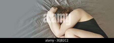 Junge kaukasische Frau in schwarzer Kleidung in fetaler Position auf dem Bett mit grauen Laken, die emotionale Belastung zeigen Stockfoto