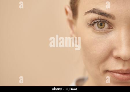 Teilgesicht einer jungen kaukasischen Frau mit fokussiertem Auge und natürlichem Make-up auf weichem beigem Hintergrund Stockfoto