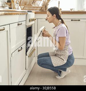 Kaukasische junge Frau, die in der Küche mit Schürze backt und vorausschauend den Ofen überprüft Stockfoto