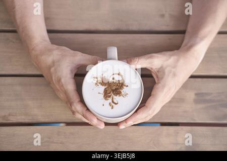 Kaukasischer Erwachsener hält Kaffeetasse mit künstlerischem Schaumdesign auf Holztisch Stockfoto