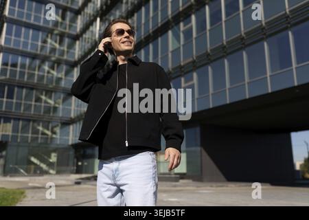 Ein junger Mann steht vor einem eleganten Glasgebäude, trägt eine Sonnenbrille und eine schwarze Jacke. Er lächelt und redet auf seinem Handy unter dem klaren Blau Stockfoto