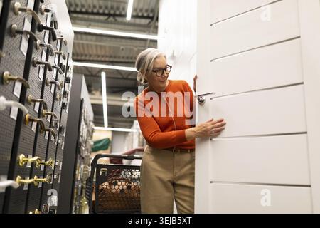 Eine Frau mit Brille und einem orangefarbenen Pullover untersucht eine weiße Tür in einem Geschäft für Heimwerker. Sie konzentriert sich auf die Merkmale der Tür in der Hardware Stockfoto