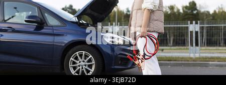 Nahaufnahme der Frau mit Kabeln zum Laden der Batterie in der Nähe des kaputten Autos mit angehobener Motorhaube Stockfoto