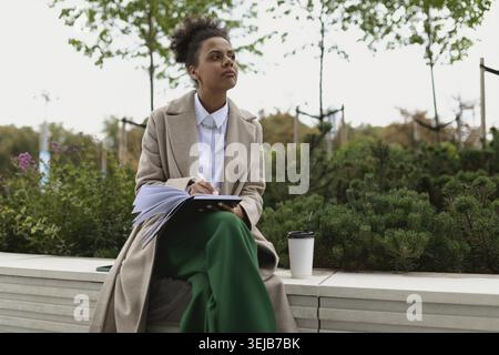 afroamerikanische Studentin macht Notizen in einem Notizbuch, während sie draußen Kaffee trinkt Stockfoto