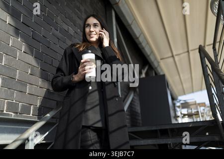 Porträt einer erfolgreichen modernen unabhängigen Frau in Geschäftskleidung, die auf einem Handy auf einer schwarzen Treppe mit einer Tasse Kaffee in der Hand spricht Stockfoto