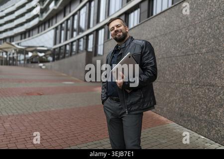 Porträt eines erwachsenen männlichen Unternehmers mit Laptop neben einem Verwaltungsgebäude in der Stadt Stockfoto