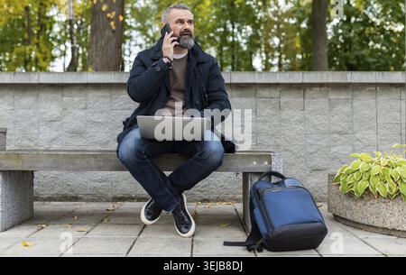 Geschäftsmann in Smart-Casual-Kleidung spricht auf einem Mobiltelefon und arbeitet online in einem Stadtpark an einem Laptop Stockfoto
