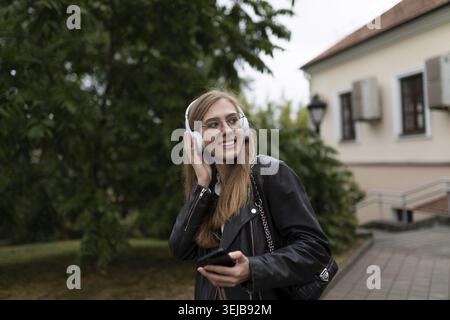 Lächelnde junge Studentin, die Musik mit Kopfhörern vor dem Hintergrund der urbanen Landschaft hört Stockfoto