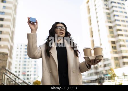 Junge, stilvolle Frau mit zwei Tassen Kaffee in den Händen, die einem Freund in der Stadt winkt Stockfoto