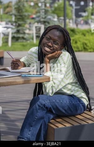 Die junge afroamerikanische Frau lächelt, während sie draußen mit einem Notizbuch, Stift und Laptop an einem sonnigen Tag studiert Stockfoto
