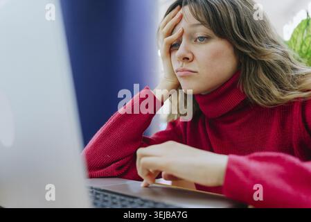 Junge Frau, die sich müde und erschöpft fühlt, während sie von ihrem Heimbüro aus arbeitet und Schwierigkeiten hat, einen anspruchsvollen Designauftrag auf ihrem Laptop zu erledigen Stockfoto
