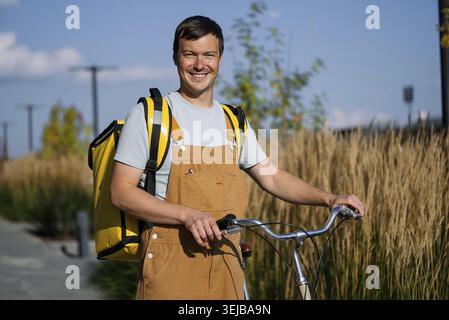 Lächelnder Mann, der als Fahrradkurier arbeitet, eine gelbe Liefertasche und einen Overall trägt, Güter mit einem Fahrrad in einem städtischen Gebiet transportiert, symbolisiert den Modus Stockfoto