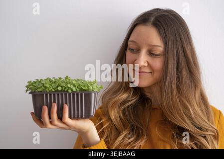 Eine zufriedene junge Frau in einer senfgelben Spitze bewundert einen Topf mit üppigen grünen Mikrogrün, die sie sanft in ihren Händen hält und ein gesundes Leben verkörpert. Stockfoto