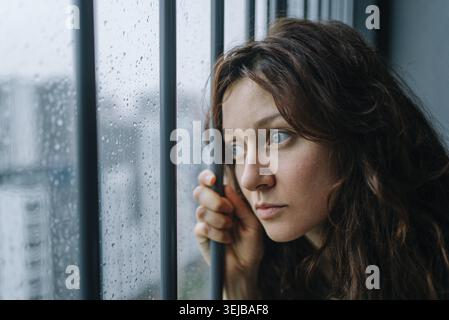 Junge Frau, die durch ein regendurchflutetes Fenster starrt, sich einsam und melancholisch fühlt, während eines Q über psychische Gesundheit, Angst und emotionale Schmerzen nachdenkt Stockfoto
