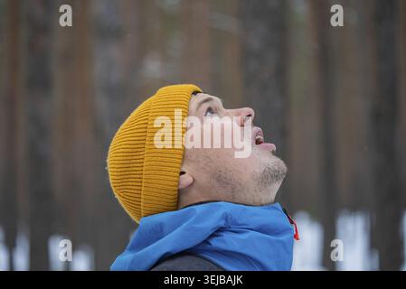 Junger Mann in gelber Mütze und blauer Jacke, der mit offenem Mund nach oben blickt, die Kälte spürt und sich wundert, während er einen leichten Schneefall in A erlebt Stockfoto