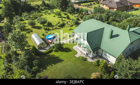 Blick aus der Vogelperspektive auf ein modernes Familienhaus, umgeben von üppigem Grün, mit einem Gewächshaus, einem kleinen Pool und gepflegten Rasenflächen Stockfoto